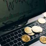 Golden Bitcoins on a laptop keyboard with a stock market chart on the screen, symbolizing cryptocurrency trading.